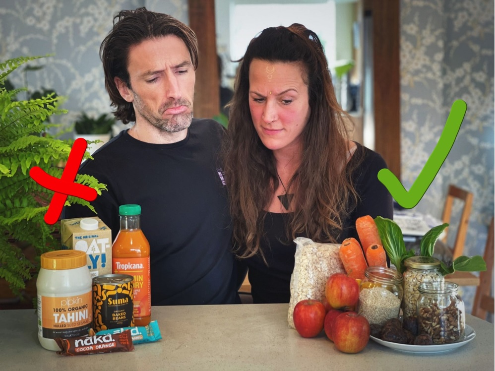 Couple stand between two piles of food, one of produce and one of products, to highlight the benefits of eating whole foods vs. processed foods